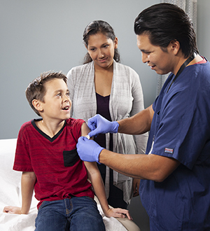 Health care provider giving boy injection in arm while parent watches.