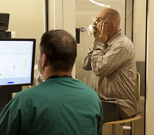 Technician performing pulmonary function tests on man in testing booth.