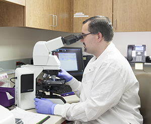 Lab technician sitting at table looking through microscope.